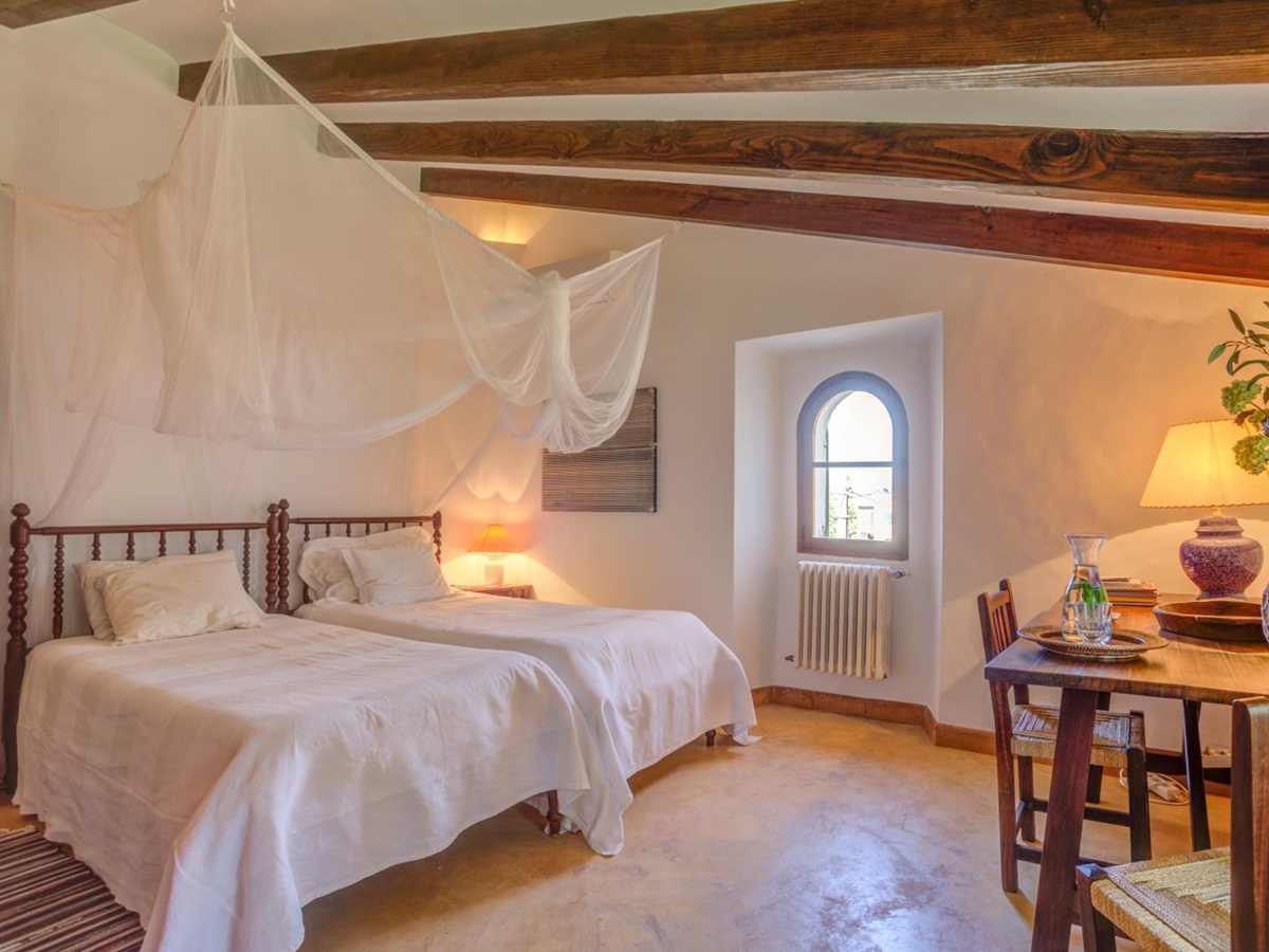 Twin bedroom with net draping, table, chairs, lamp, small window and ceiling beams at Can Canals in Mallorca, Spain