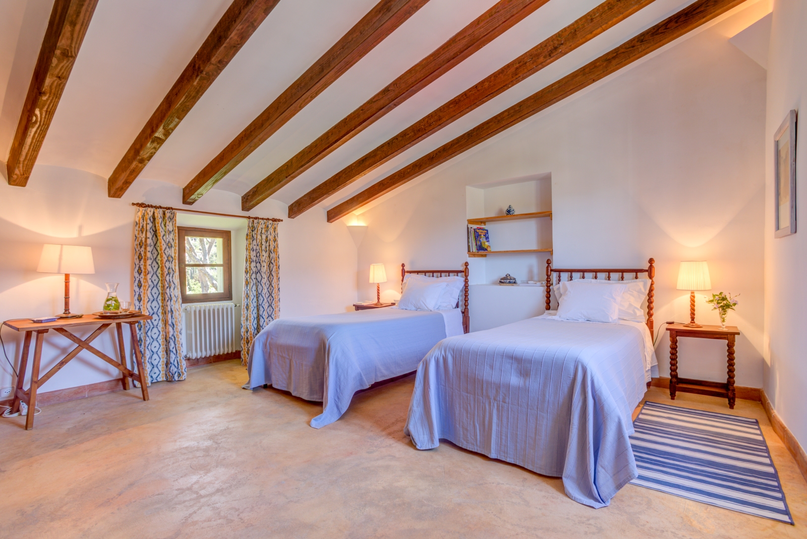 Twin bedroom with tables, lamps, beams, shelves, blue and yellow curtains and a small window at Can Canals in Mallorca, Spain