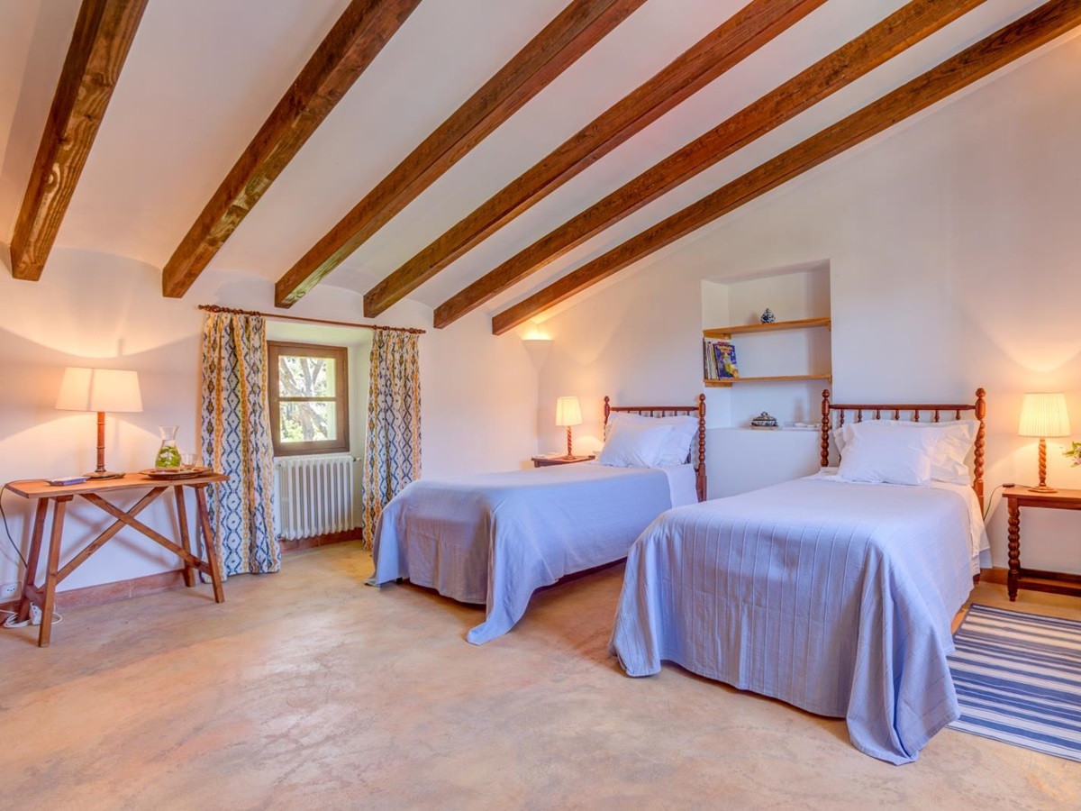Twin bedroom with tables, lamps, beams, shelves, blue and yellow curtains and a small window at Can Canals in Mallorca, Spain