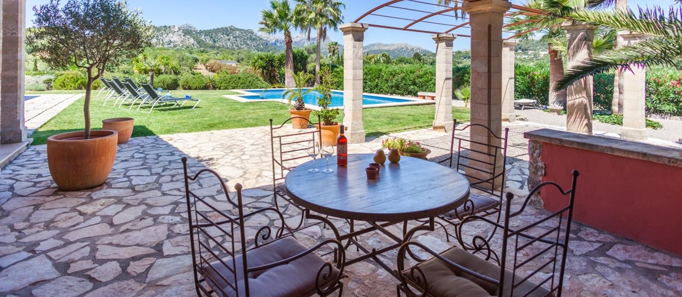 Covered outdoor dining area with round table, chairs and view of garden, pool and mountains at Can Suelo in Mallorca, Spain