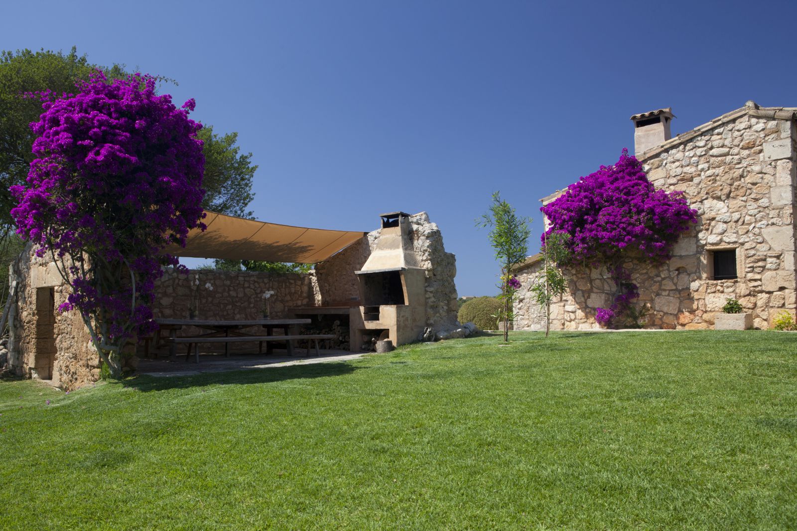 Side of stone house covered in purple flowers and long wooden table in BBQ area at villa San Lorenzo in Mallorca