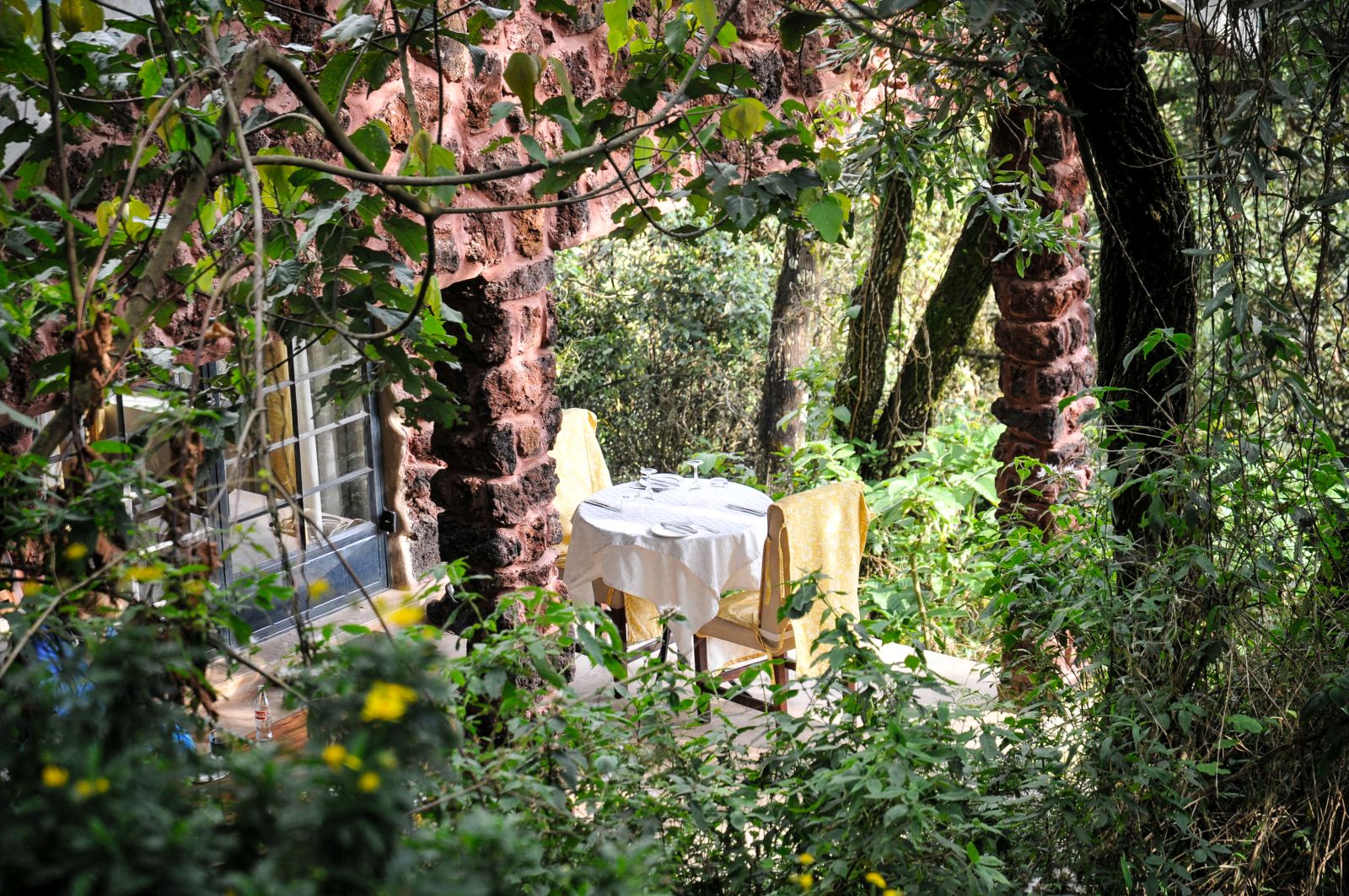 A table with white table cloth and two yellow chairs on a patio surrounded by gardens and trees at Clouds Mountain Gorilla Lodge in Uganda