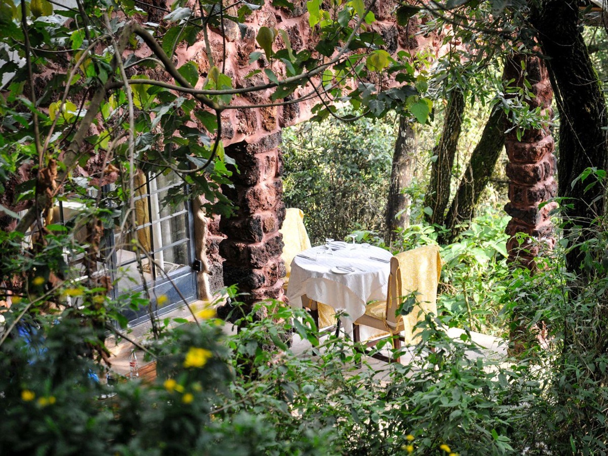 A table with white table cloth and two yellow chairs on a patio surrounded by gardens and trees at Clouds Mountain Gorilla Lodge in Uganda