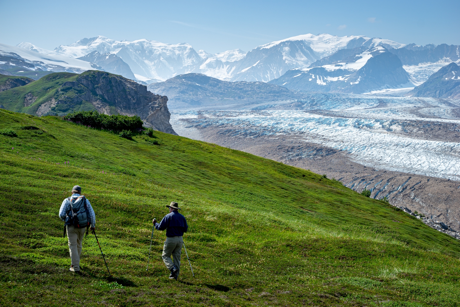 Two hikers walk parallel to a glacier
