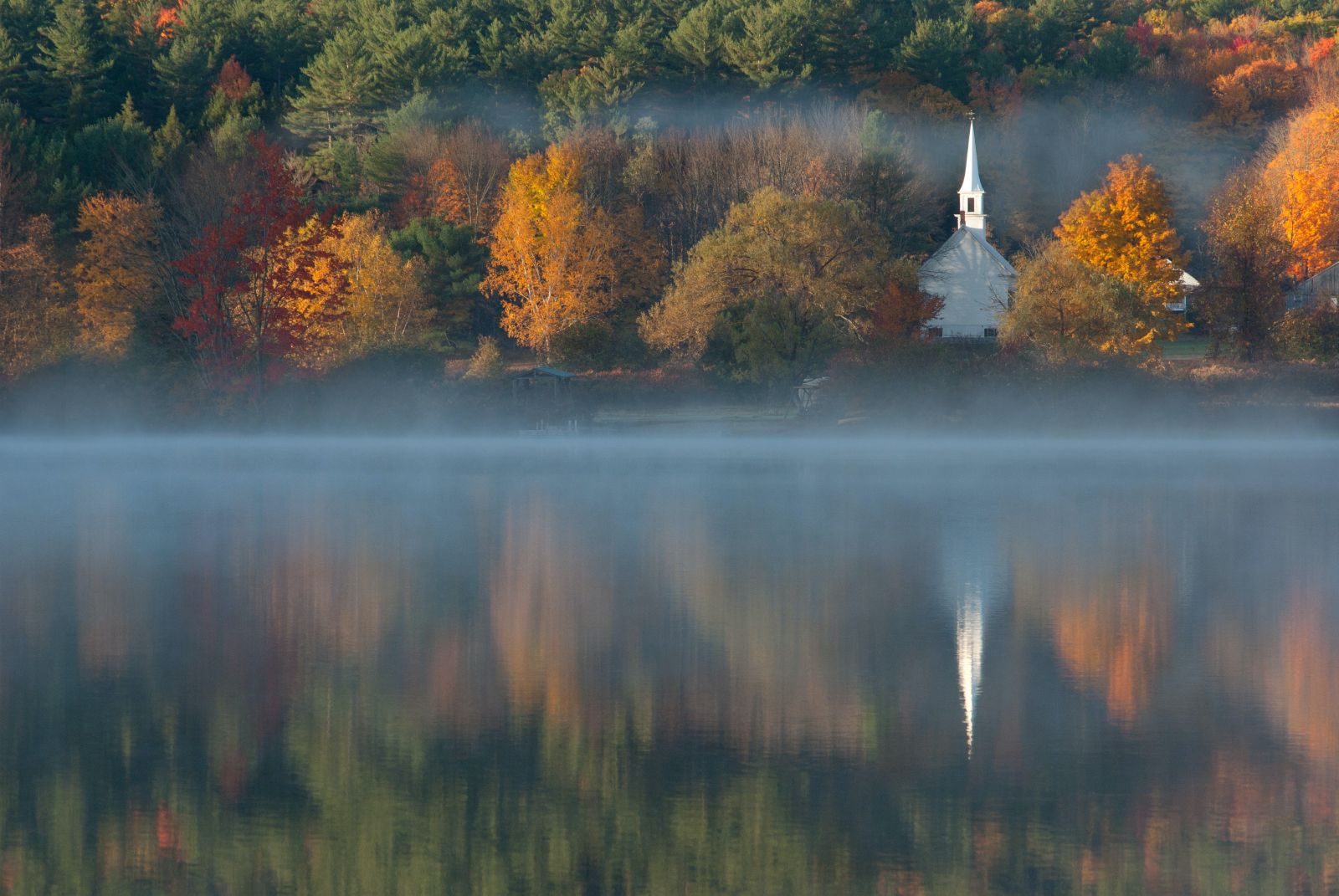 Mist rising on a lake in New Hampshire with small chapel and fall leaves