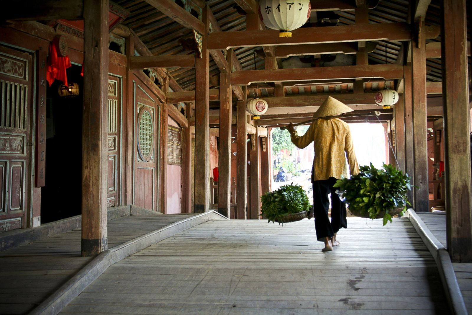 A person carrying two baskets of green vegetables walking over a Japanese covered bridge in Hoi An