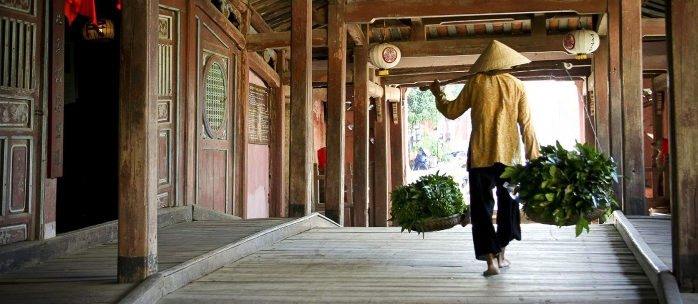 A person carrying two baskets of green vegetables walking over a Japanese covered bridge in Hoi An