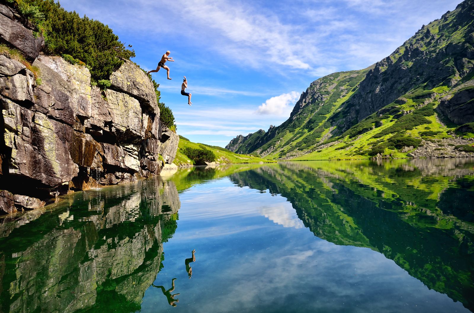 A couple jumping into a clear lake with mountain reflection